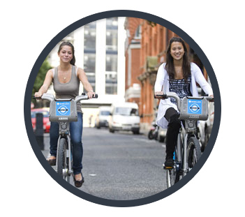 Two smiling girls riding Barclays bikes side by side on the streets of London.