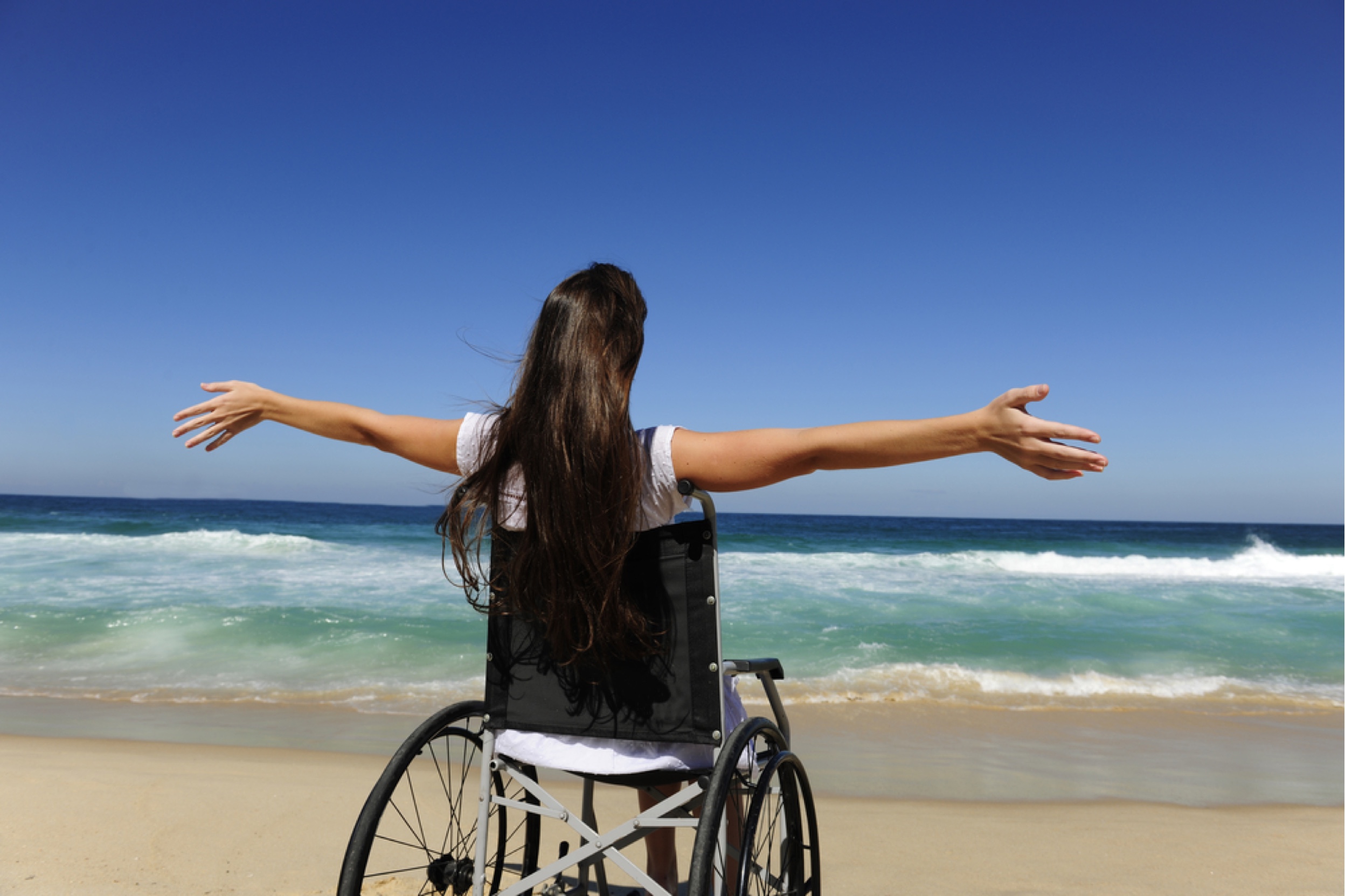 Woman in wheelchair holding arms in the air looking out at the water.