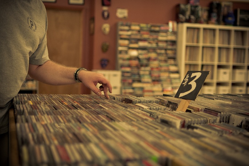 Shows a photo of a man's arm inside a record store, browsing through a pile of records marked for $3.